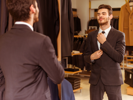Modern young handsome businessman dressed in classical suit adjusting a tie in front of the mirror while standing in the suit shop